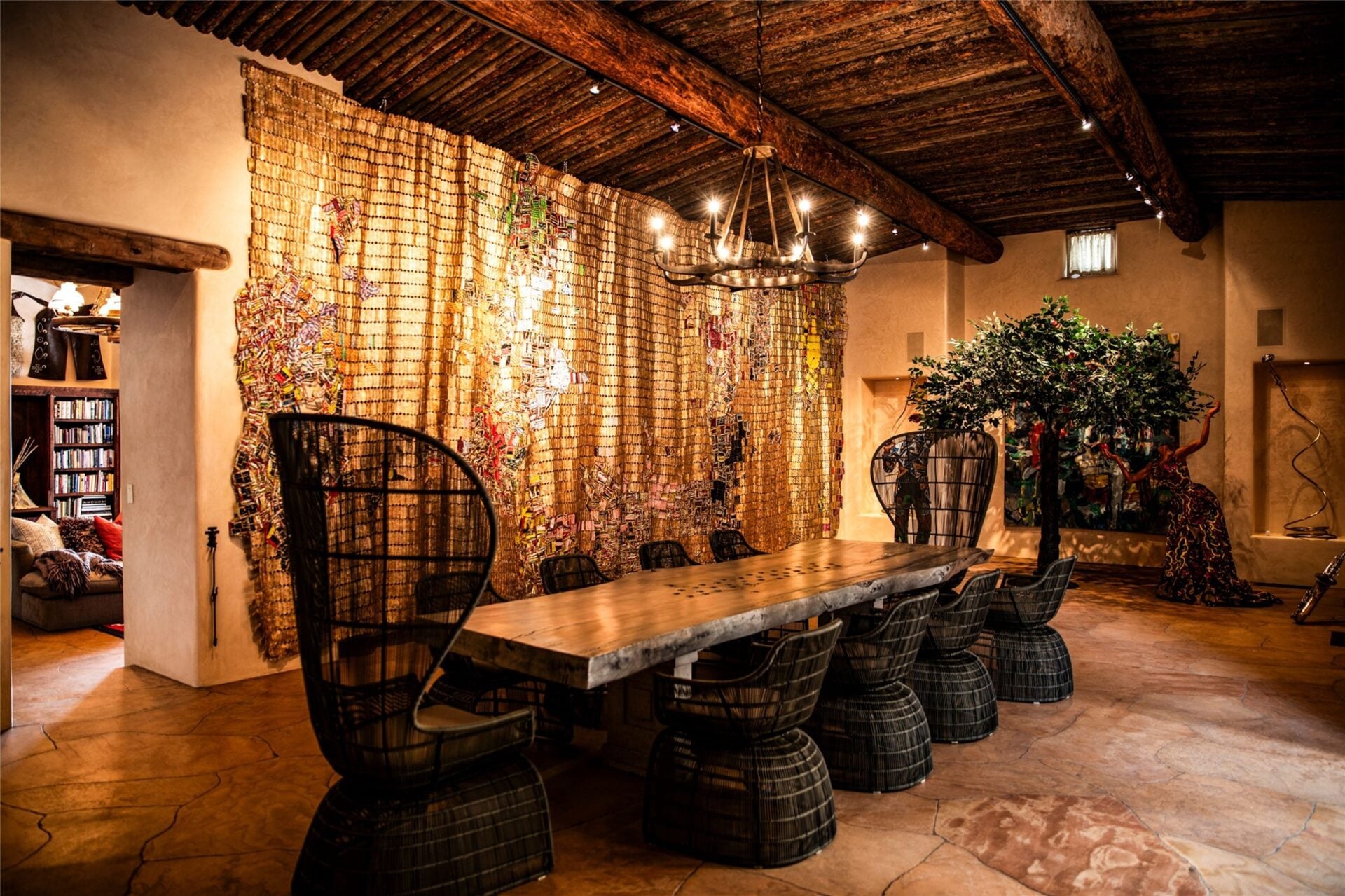 A long live-edge table runs through the center beneath an iron ring chandelier, with woven sculptural chairs lining both sides and taller wingback seats marking the ends.