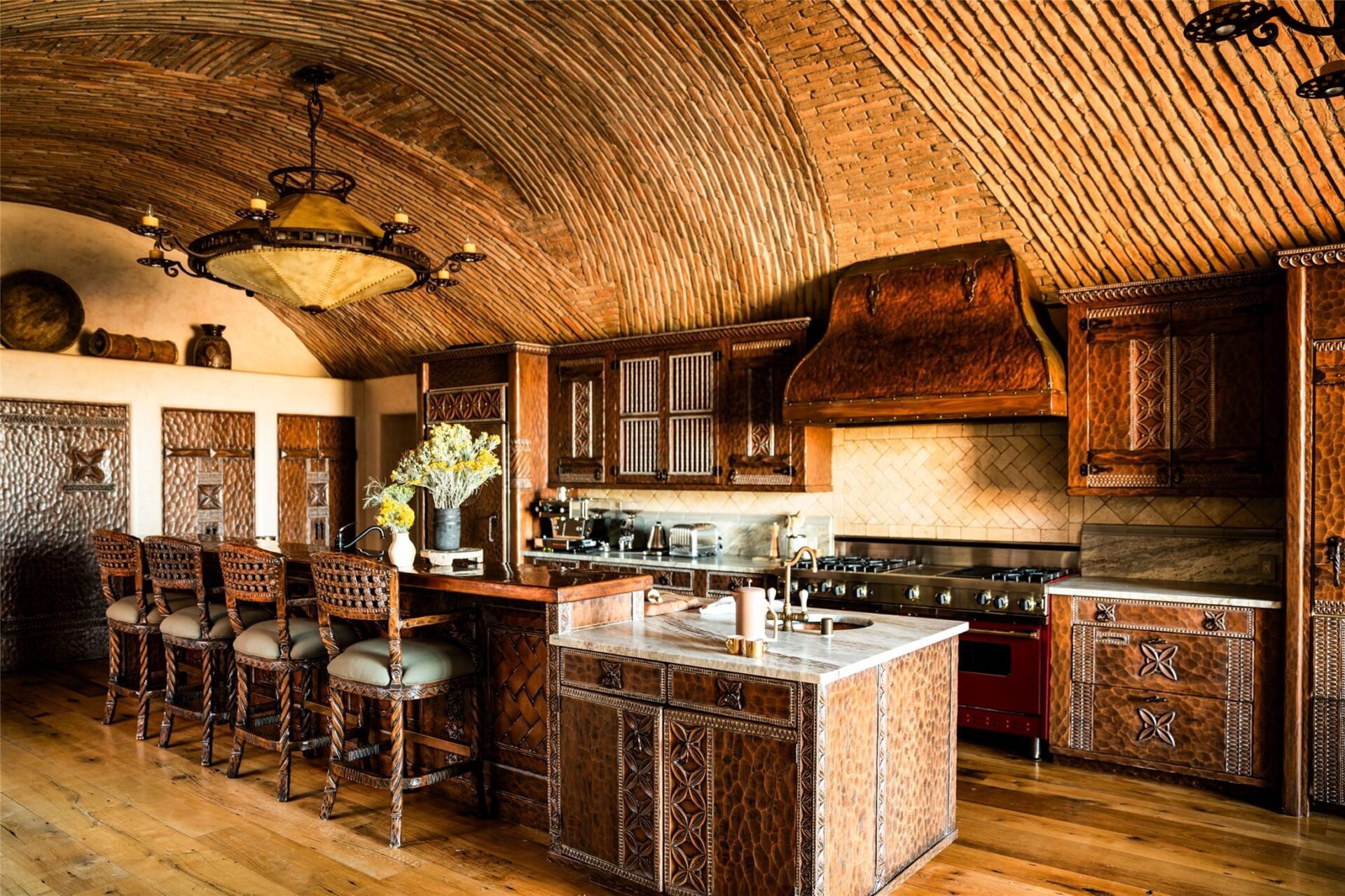 Deeply carved wood cabinetry wraps the perimeter, while hammered copper panels, carved drawers, and a large copper range hood give the room a dense, highly detailed finish.