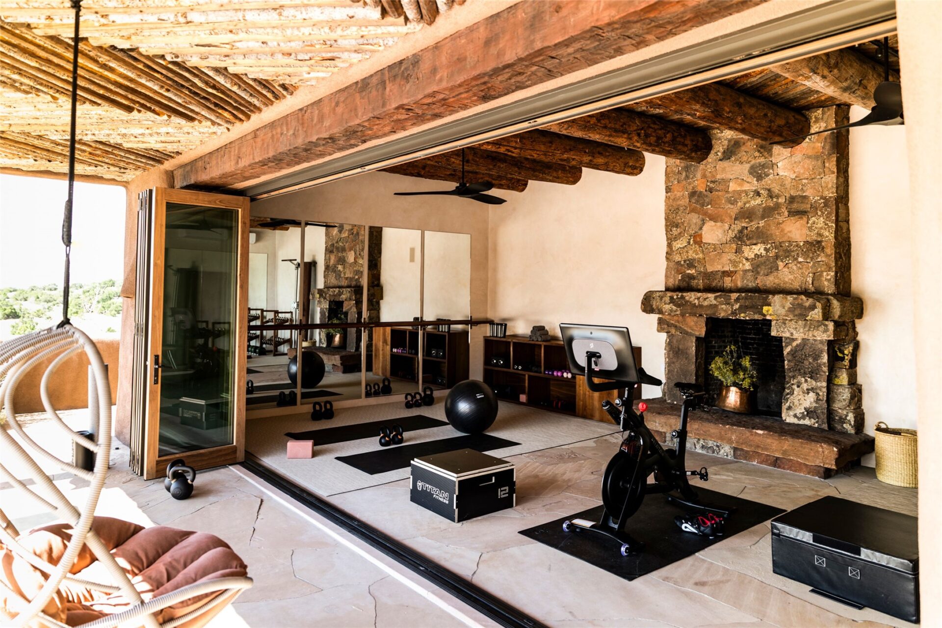 A detached building opens into a private gym through a wall of folding glass doors with mirrored walls and a large stacked-stone fireplace. 