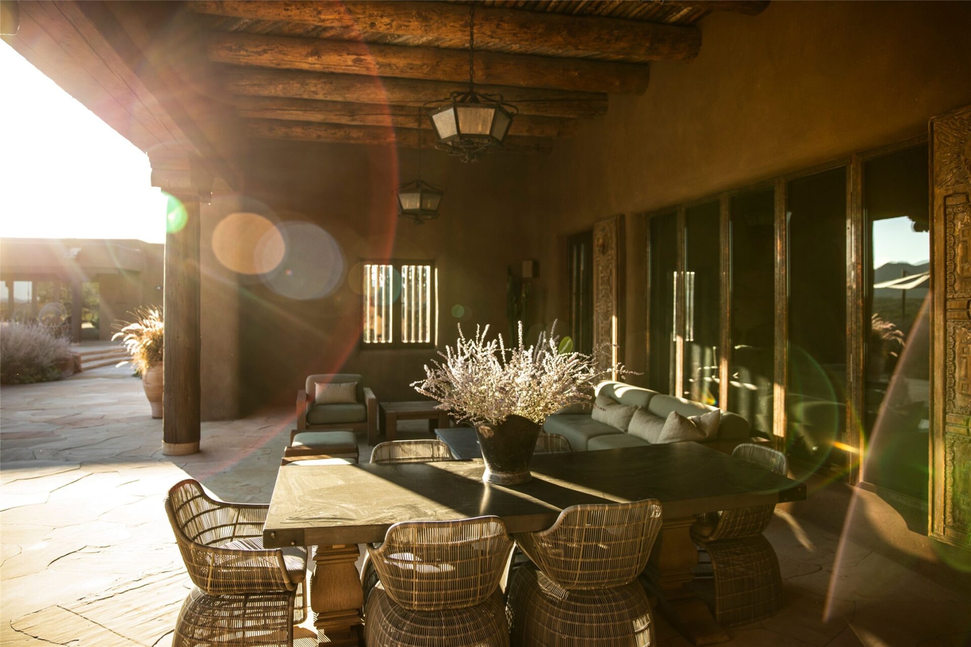 A covered dining area sits along the pool deck with a long table filling the center of the shaded terrace, with a nearby sofa and lounge chairs creating a separate seating area beside the glass doors of the hacienda.