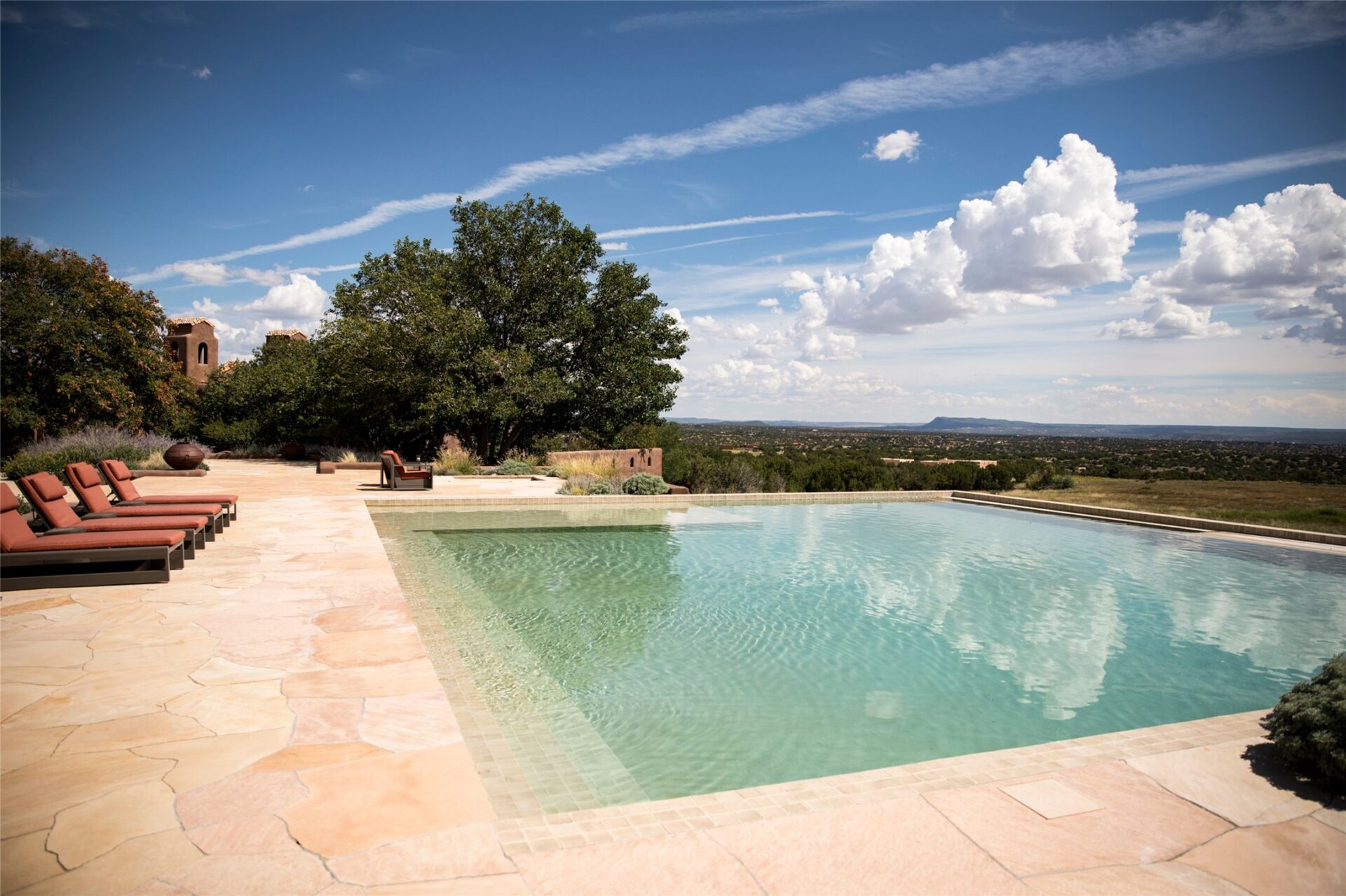 A wide stone terrace wraps the water with rows of lounge chairs arranged along one side, while low adobe walls and native plants blend the pool into the surrounding terrain.