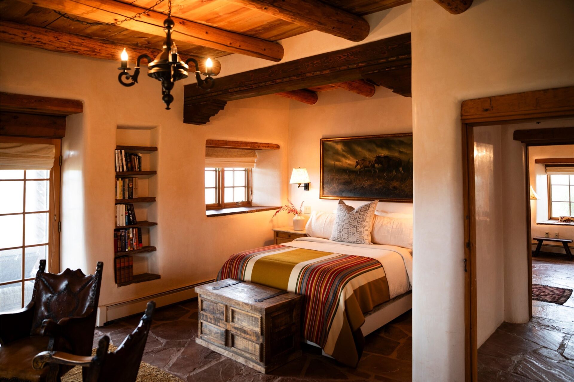 The primary suite includes a bed placed beside recessed shelving and small punched windows, while a sitting space occupies the front of the room near glass doors.