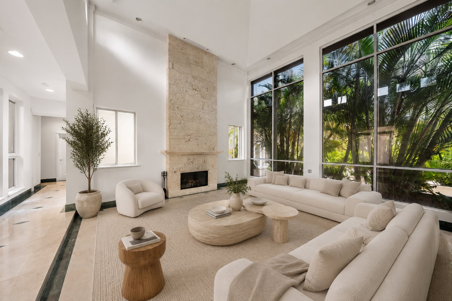 A floor-to-ceiling stone fireplace anchors one wall, while the opposite side is lined with expansive windows that overlook the surrounding tropical landscaping. 