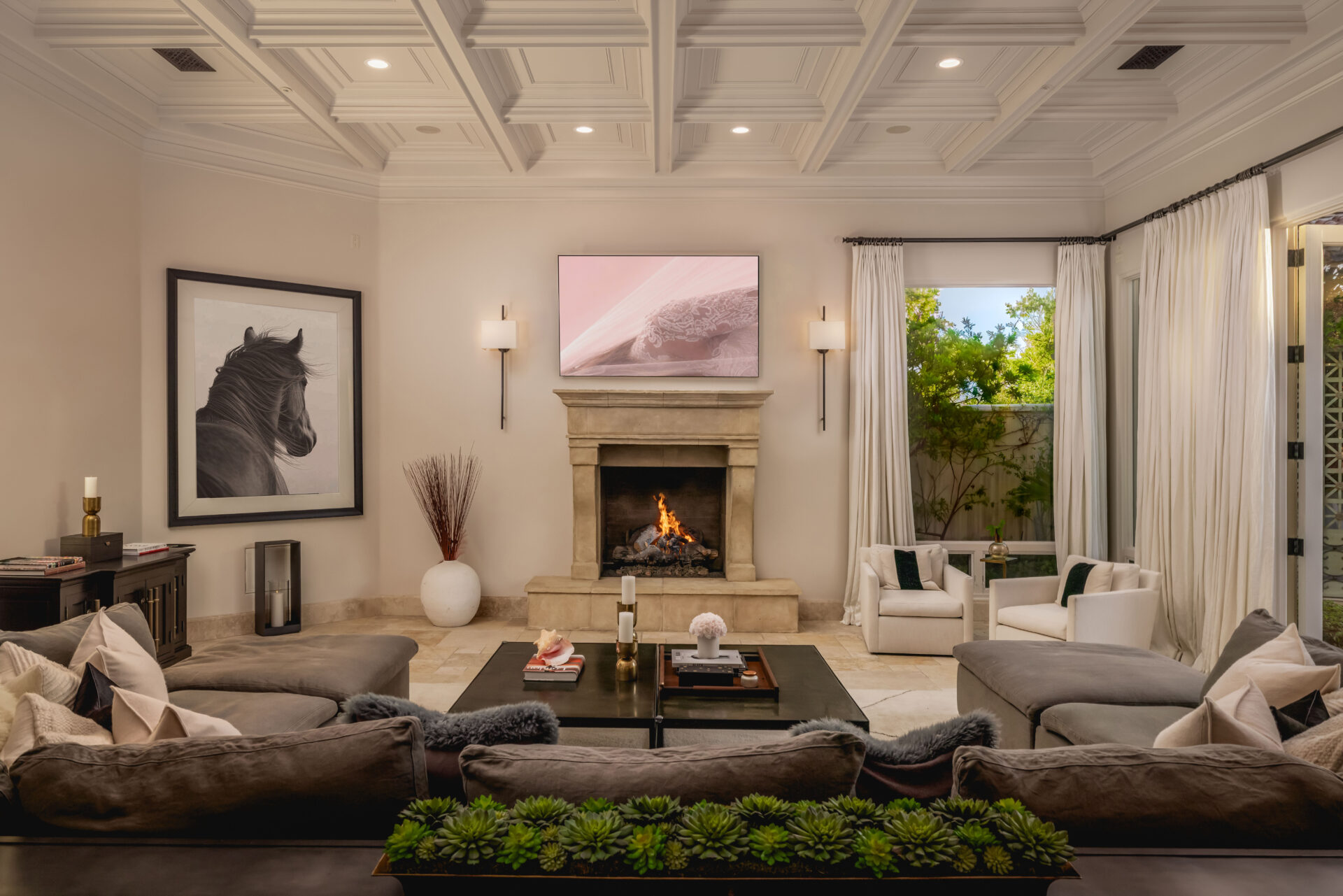 A large gray sectional curves around a pair of low square coffee tables, directing the room toward a limestone fireplace with a television mounted above and slim wall sconces on either side. 