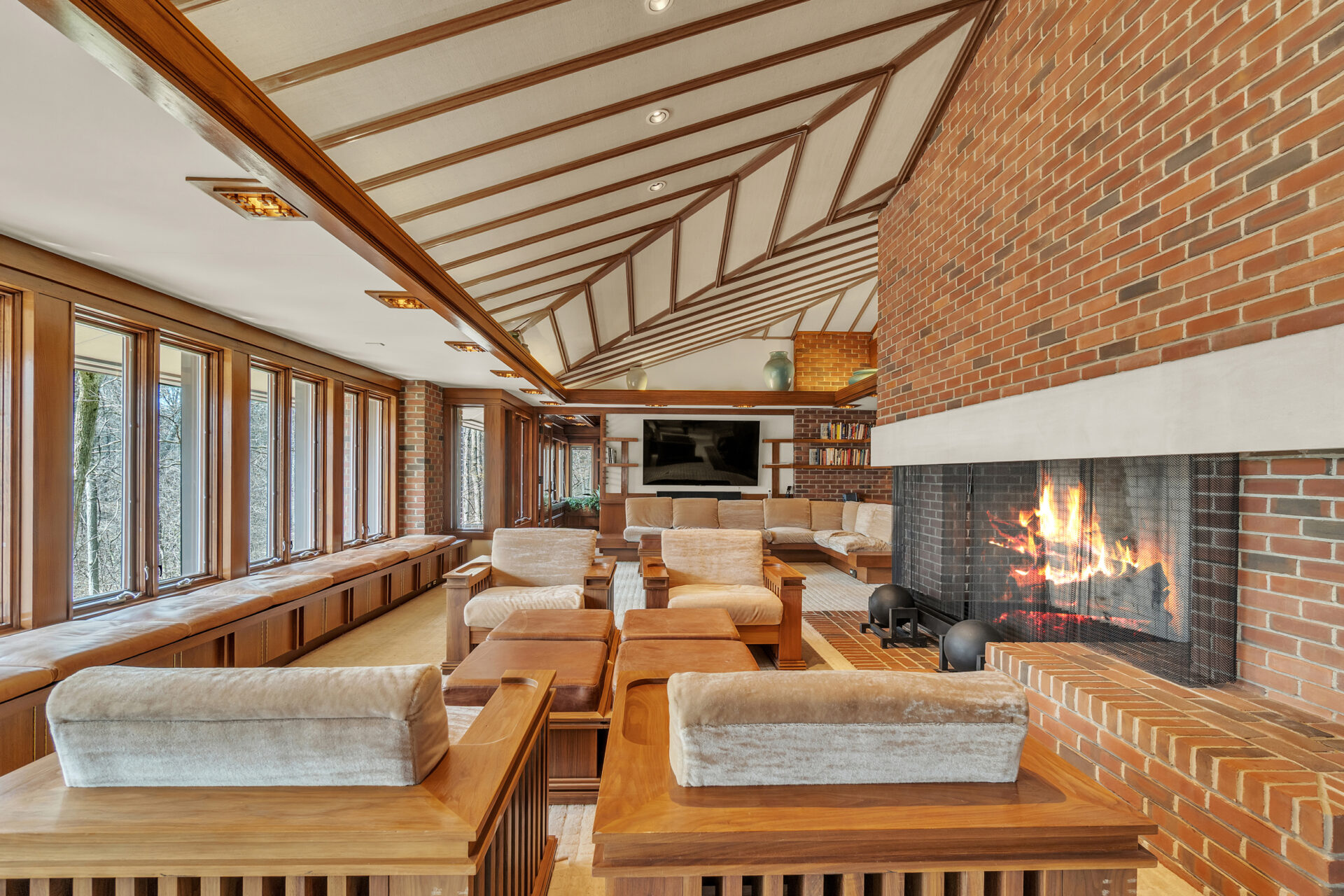 Surrounding it are built-in wood-framed lounge chairs, custom shelving, a media wall, and a grand piano, while continuous window seating stretches beneath a wall of floor-to-ceiling windows overlooking the grounds.