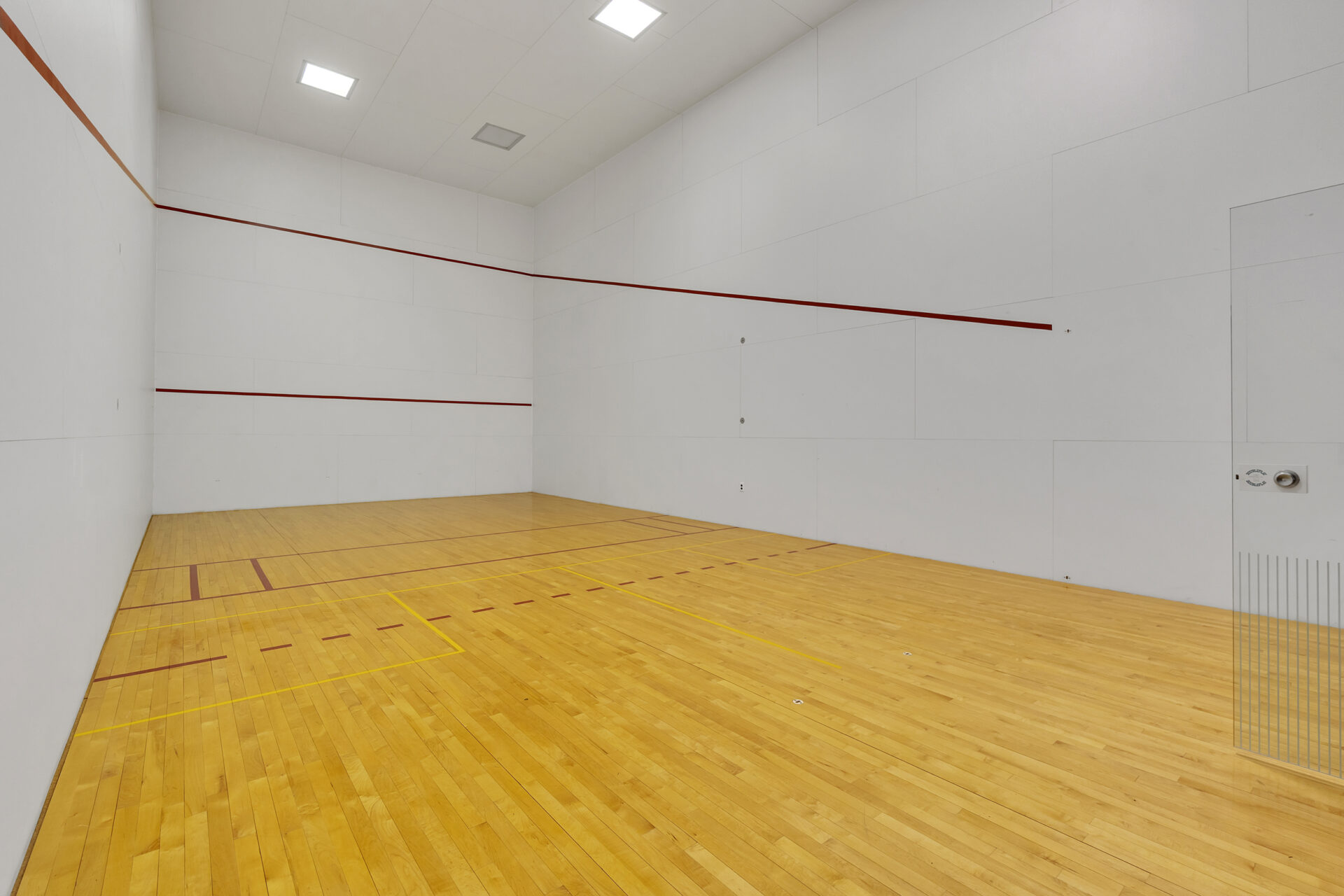 A full-size indoor racquetball court with regulation wood flooring, glass entry doors, and marked playing lines.
