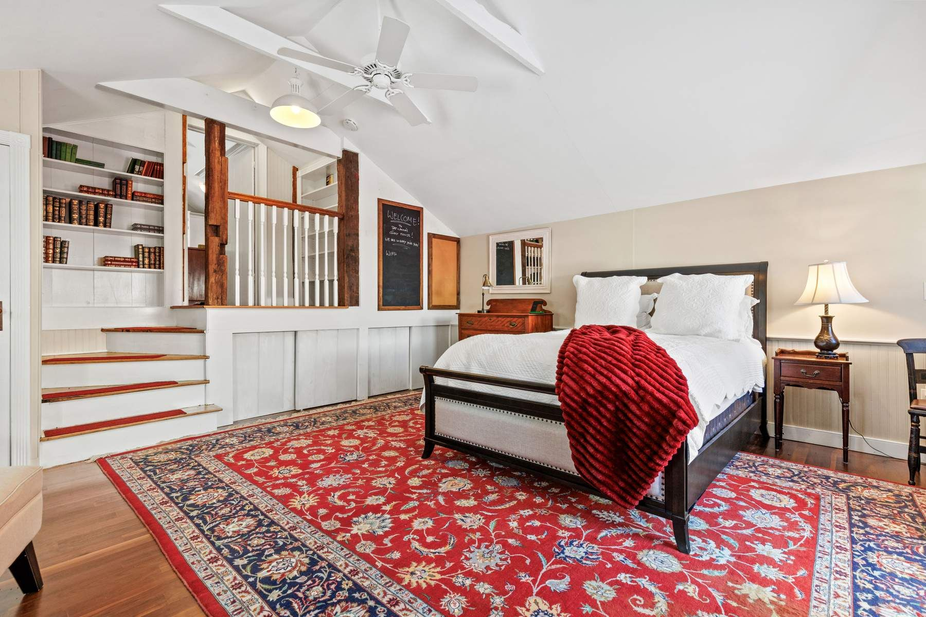 Just beyond, the bedroom occupies its own private corner of the cottage, featuring a vaulted ceiling, built-in bookshelves, and warm hardwood floors beneath a large patterned rug.