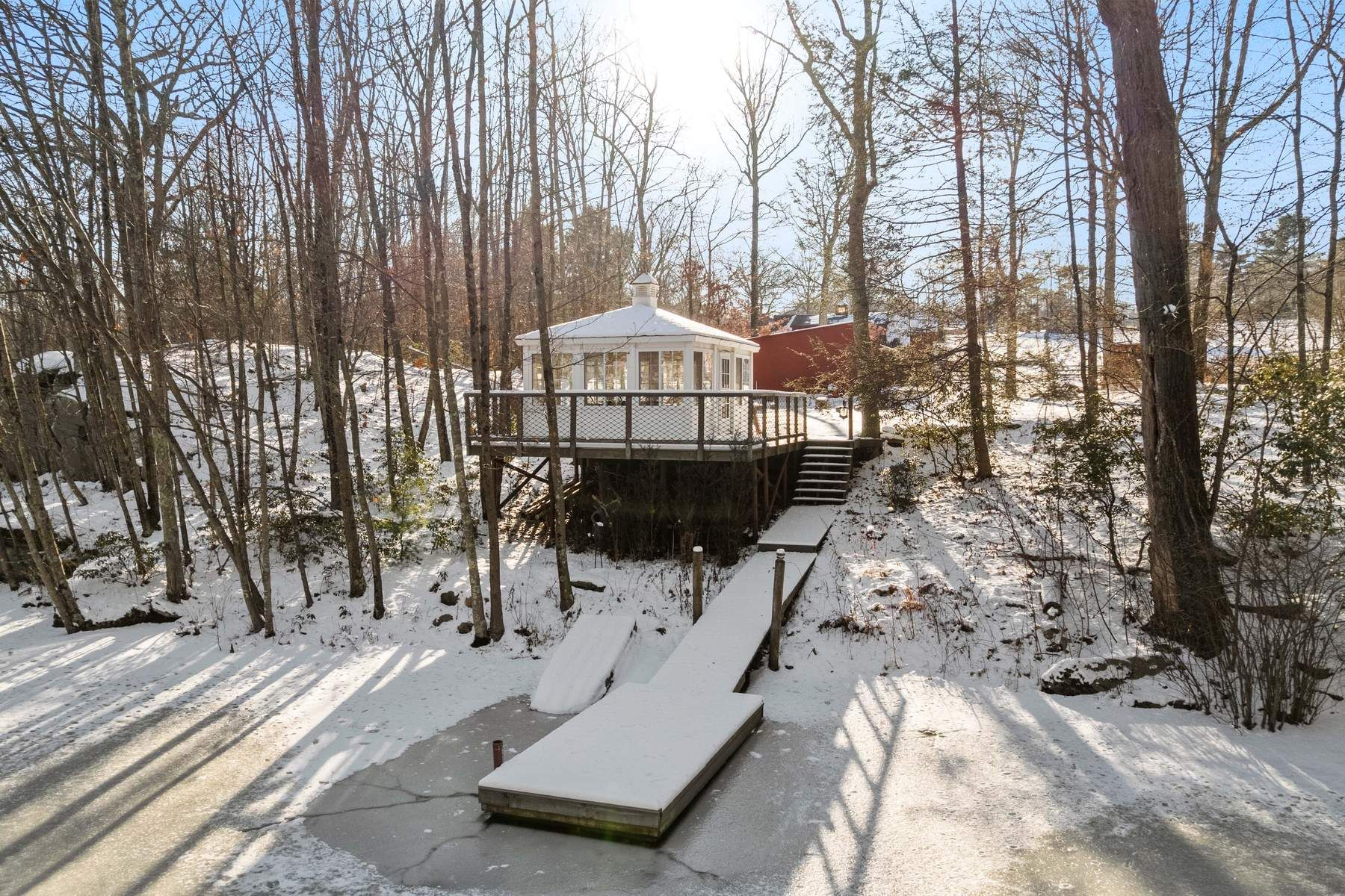 A white pond house sits on an elevated deck overlooking the water with its own dock extending to the shoreline.