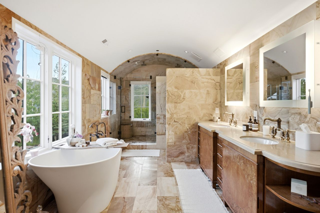 Bathroom finished almost entirely in warm-toned stone, with dual vanities stretching along one wall and a freestanding soaking tub positioned beneath oversized windows. 
