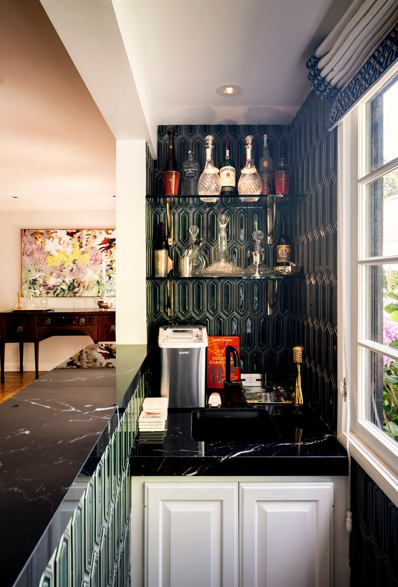 Black marble countertops are paired with geometric emerald-green tile that wraps the walls and bar front, while floating glass shelves display crystal decanters and spirits.