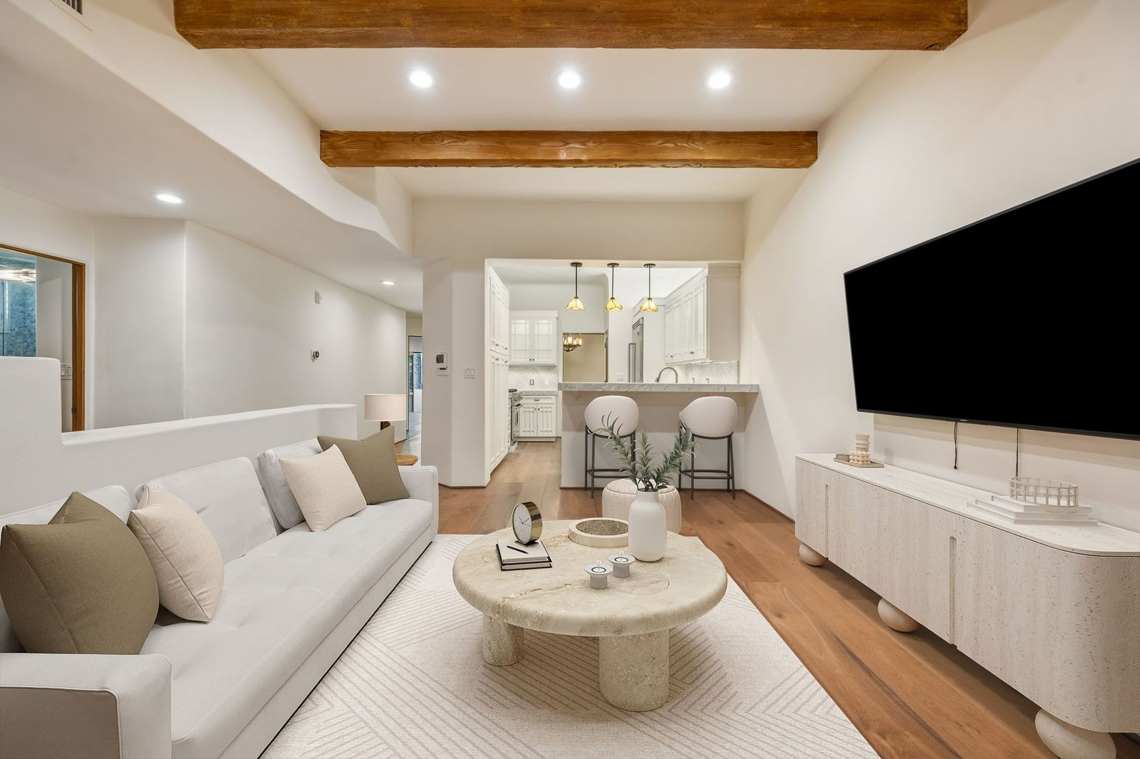 Exposed wood ceiling beams contrast with the light walls and wide-plank hardwood floors, while a soft neutral palette and rounded stone coffee table give the space a clean, contemporary feel. 