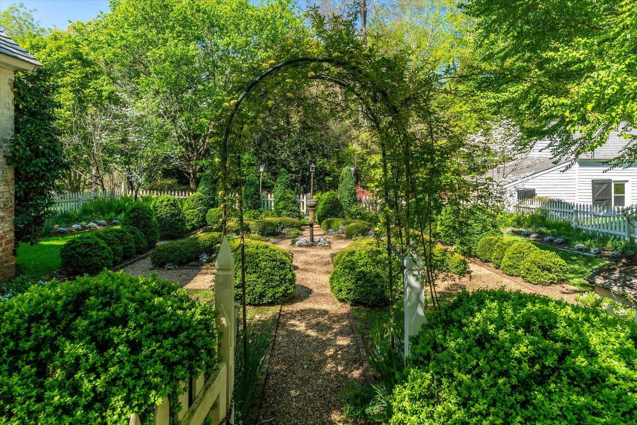 A formal boxwood garden unfolds with clipped hedges, gravel walkways, and a stone urn serving as its centerpiece.
