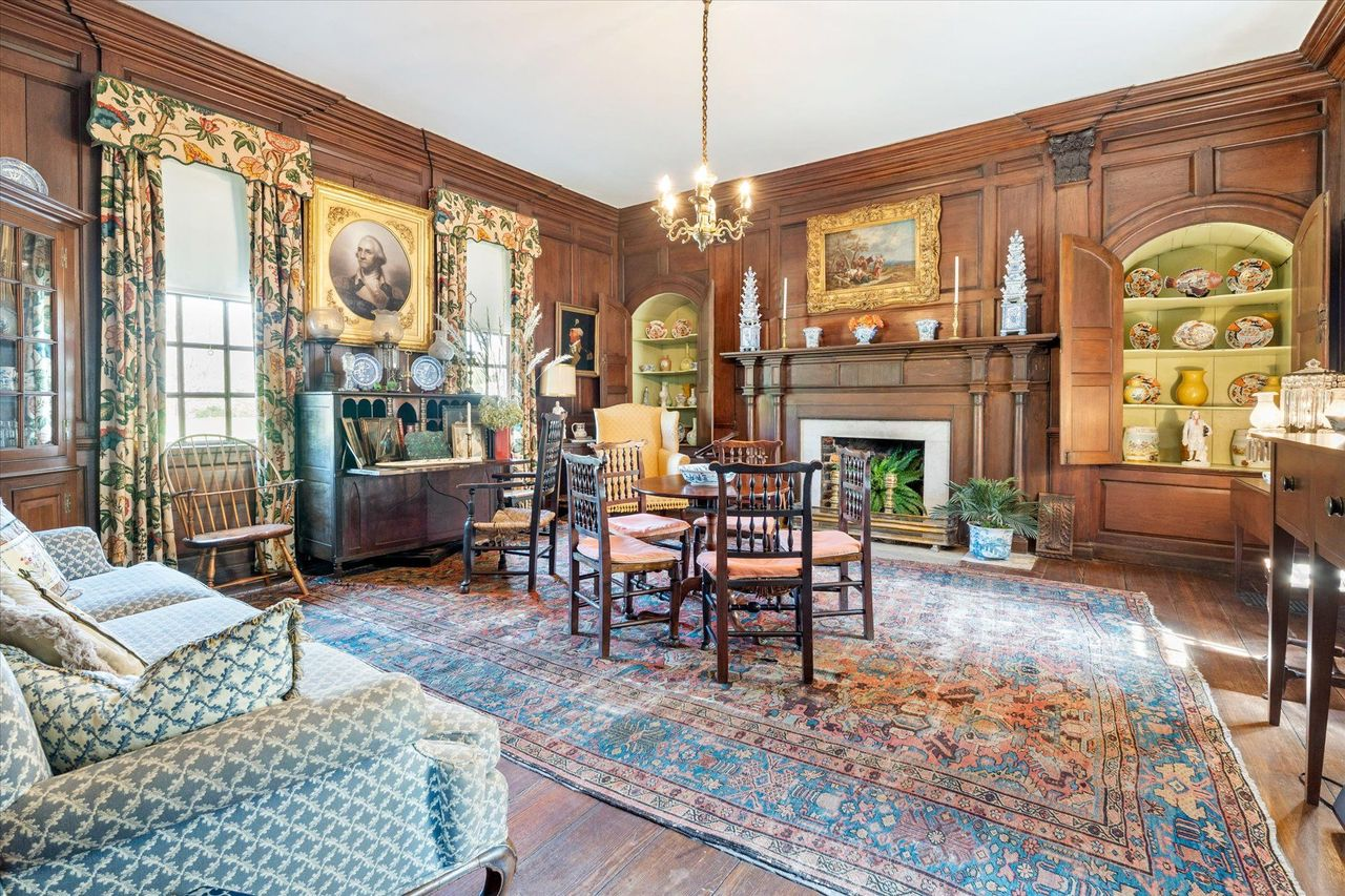 A fireplace sits at one end of the room beneath historic artwork, while built-in display alcoves, antique furniture, and a large Persian rug.