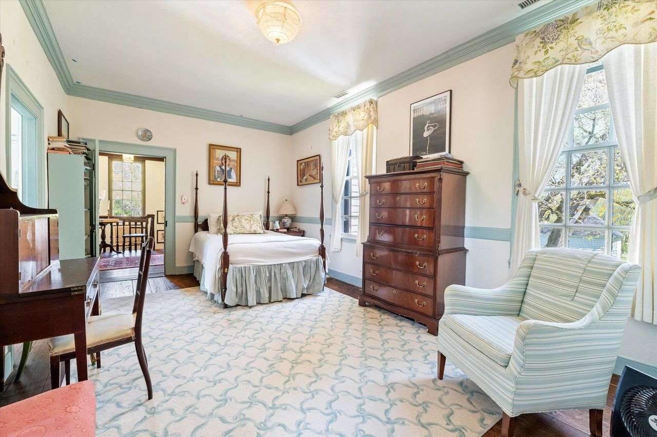 A smaller guest bedroom provides a space with soft painted trim, wide-plank floors, and tall period windows overlooking the grounds.