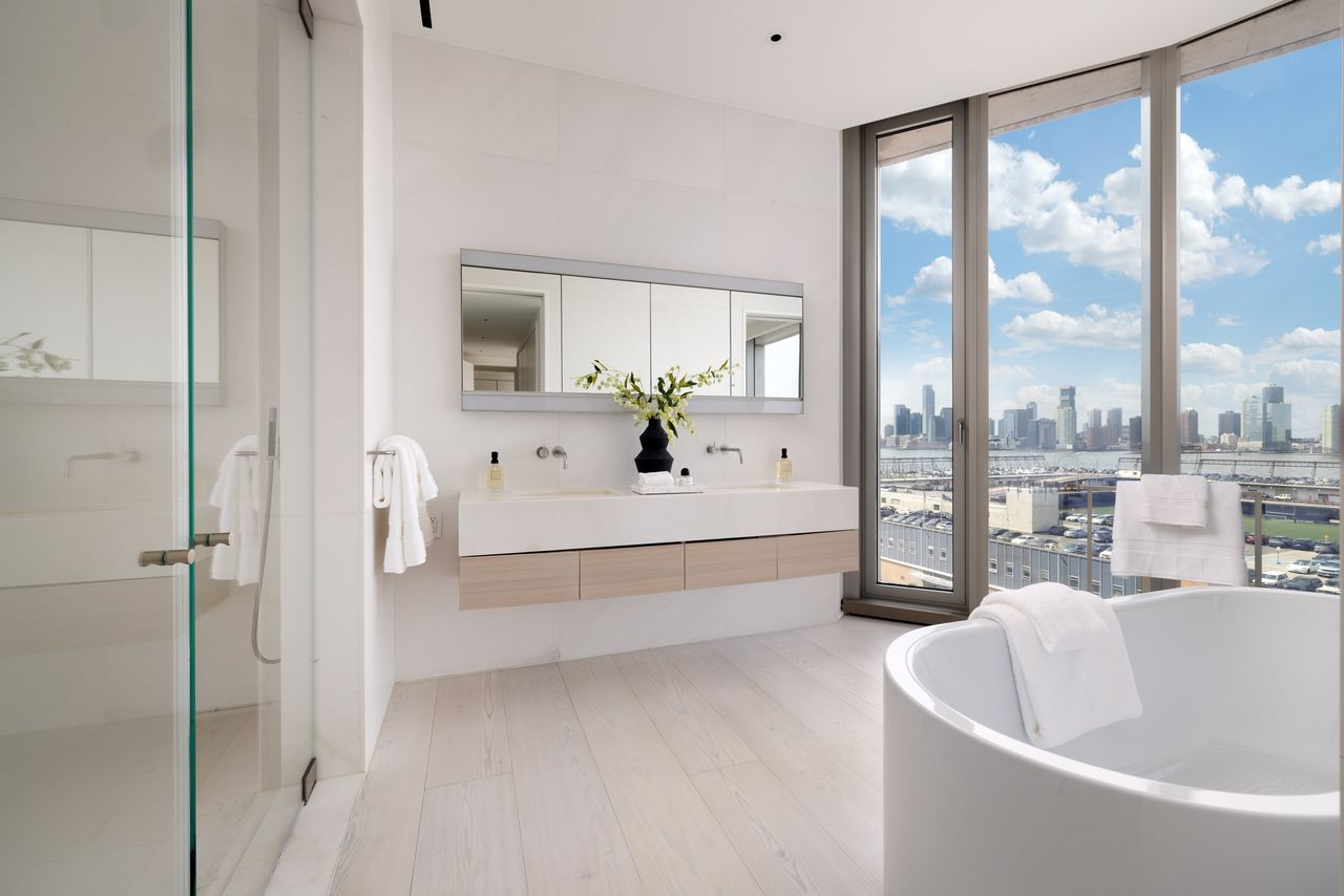 A floating double vanity and a freestanding soaking tub are positioned beside the windows, while a glass-enclosed shower sits across from them.
