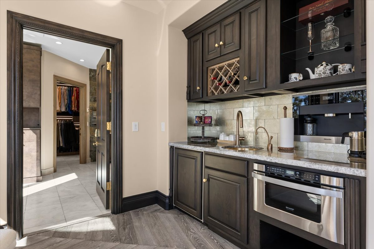 A fully equipped wet bar with dark custom cabinetry paired with stone countertops, a built-in Miele coffee station, wine storage, a sink, and open display shelving for glassware and spirits.