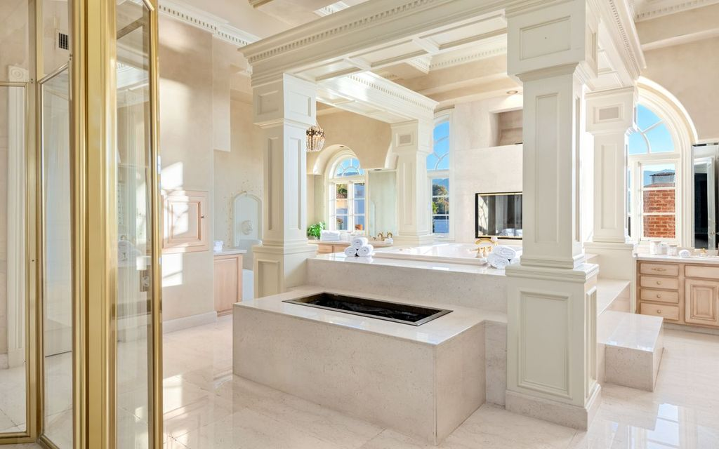 A large soaking tub sits at the center of the room, complemented by expansive vanities on either side, a glass-enclosed shower, and generous dressing space.
