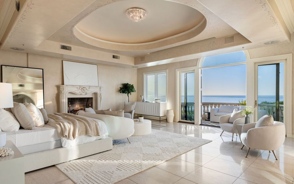 Bedroom is centered around a carved stone fireplace, with a separate sitting area positioned beside French doors that open to a balcony. 