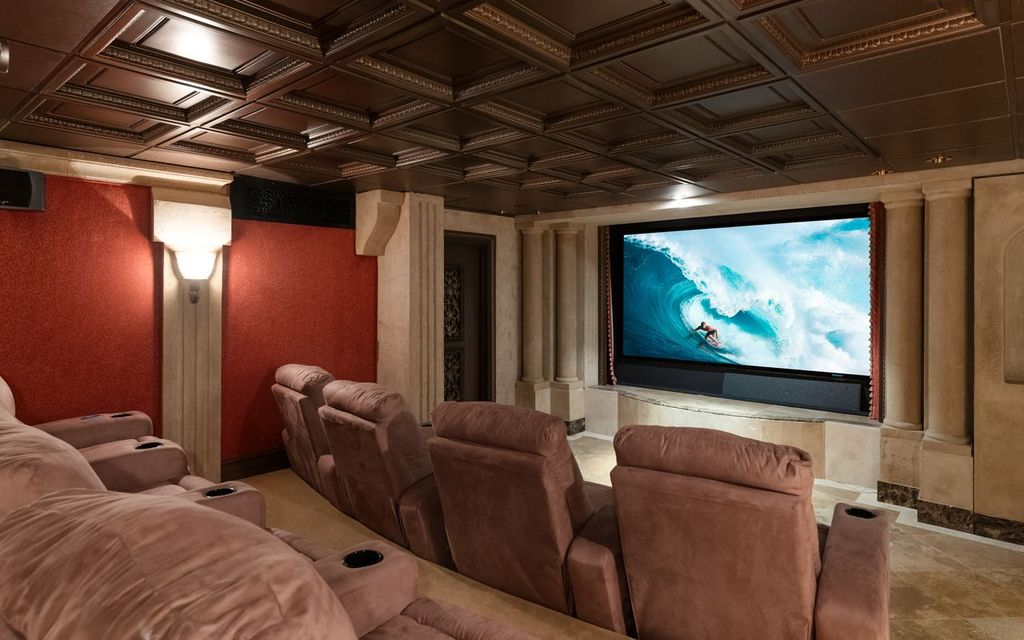 The home theater is finished with coffered ceilings, decorative columns, and tiered reclining seating arranged around a large projection screen.