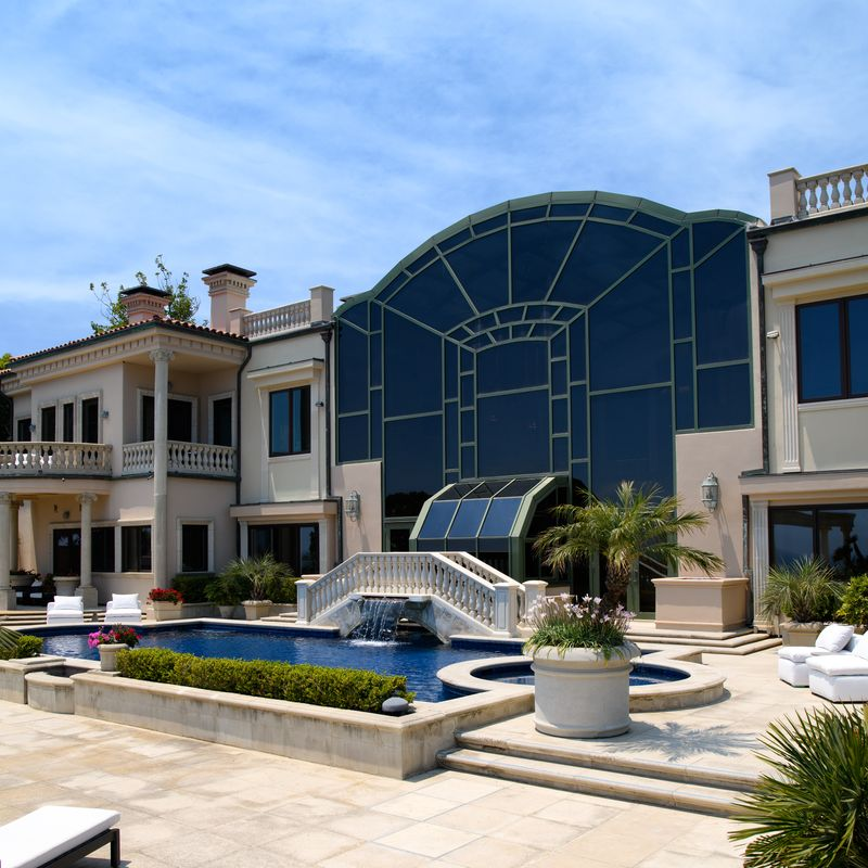 Wide terraces surround the pool with rows of chaise lounges, while a bridge and cascading water feature connect the home's glass exterior to the outdoor entertaining spaces.