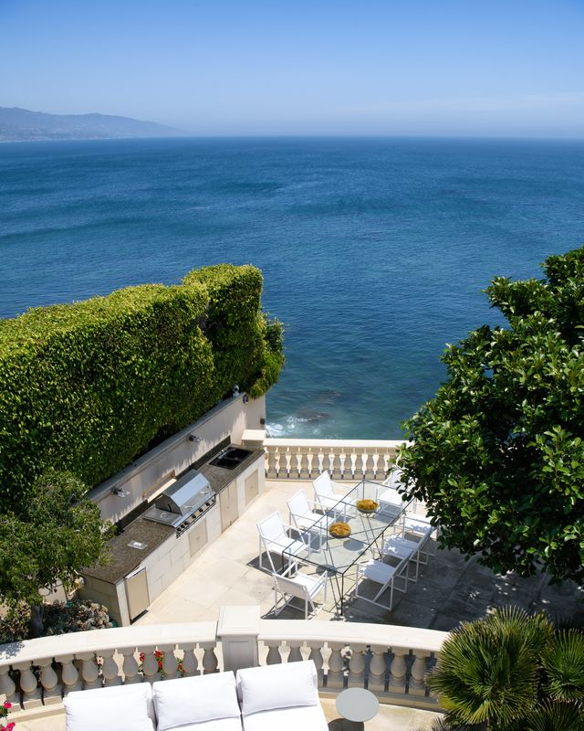 An oversized dining table sits beside a full built-in barbecue kitchen with stone countertops, creating a spot for alfresco meals while overlooking the ocean.