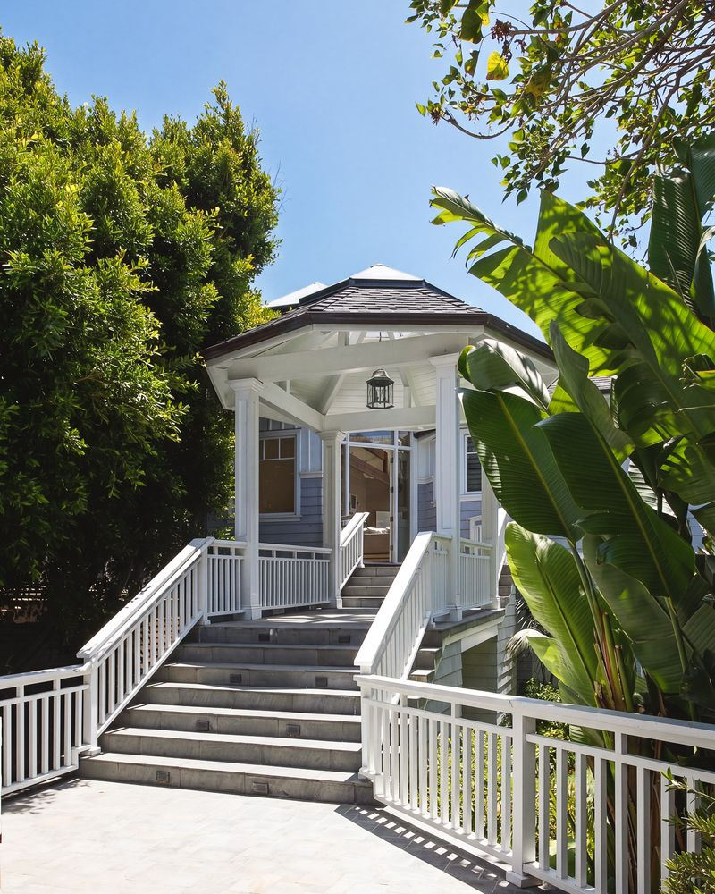 A wide staircase climbs through tropical landscaping to a covered, gazebo-style entry pavilion, where white railings, a vaulted roof, and lantern lighting create a welcoming arrival.