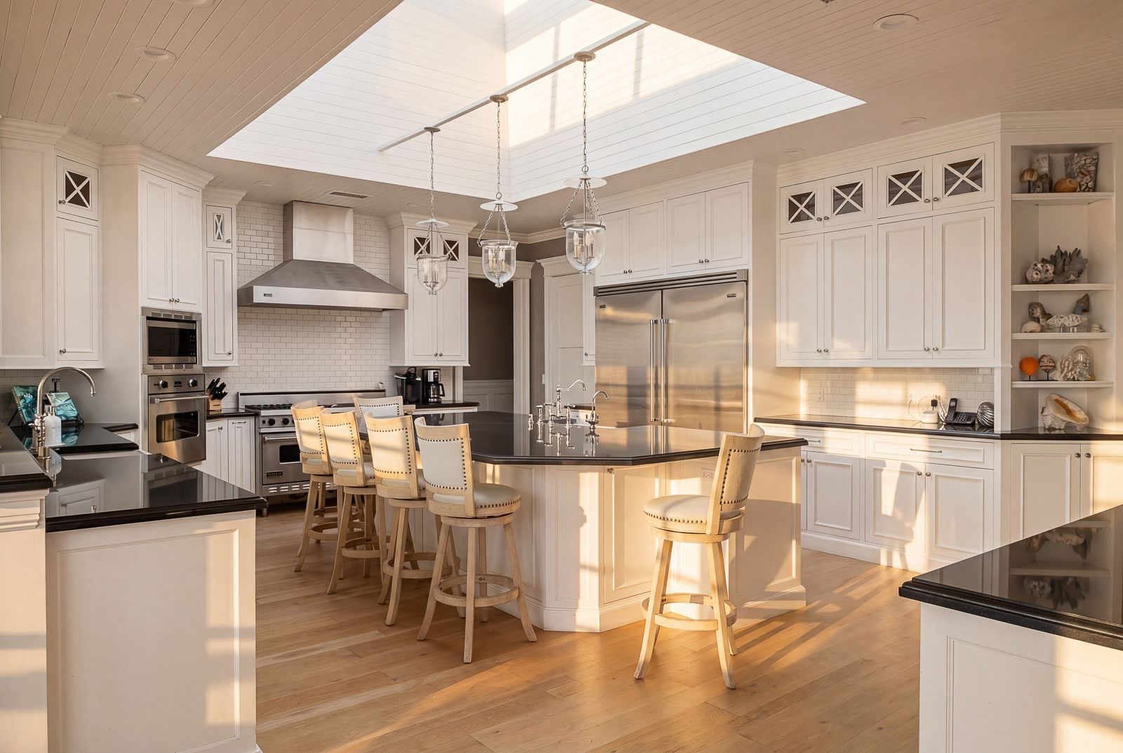 The kitchen features an oversized granite-topped island beneath a large skylight, while white cabinetry, black stone countertops, and warm wood floors create a bright coastal feel. 