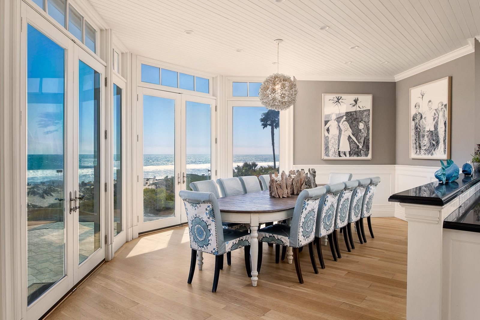 An oval dining table sits beneath a sculptural pendant light, while French doors and tall windows wrap the space with light and views of the Pacific.