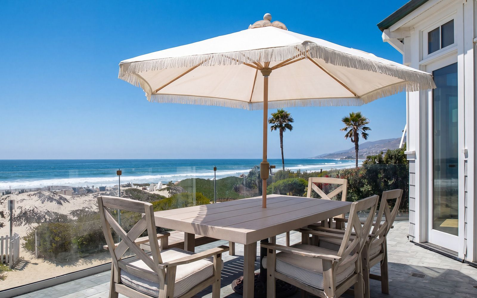 A large table sits beneath a fringed umbrella, creating a comfortable place for outdoor meals against glass railings that preserve the view.