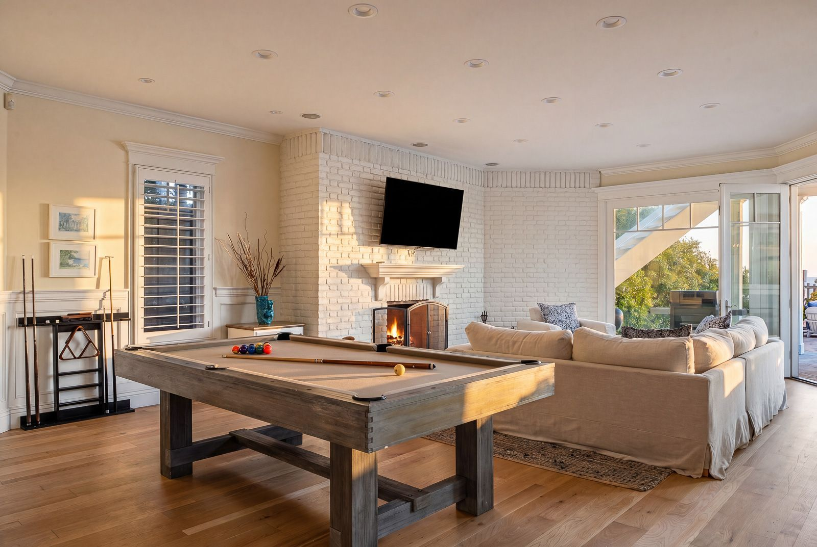 A full-size pool table is placed in the center of the room beside a white-painted brick fireplace lounge and the backyard patio doors.