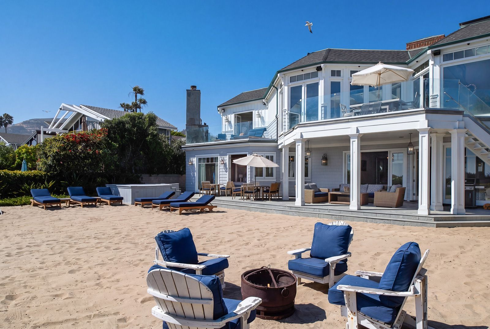 The sandy backyard is designed for spending entire days outdoors, with lounge chairs, a spa, and a fire pit arranged across the private beachfront setting. 
