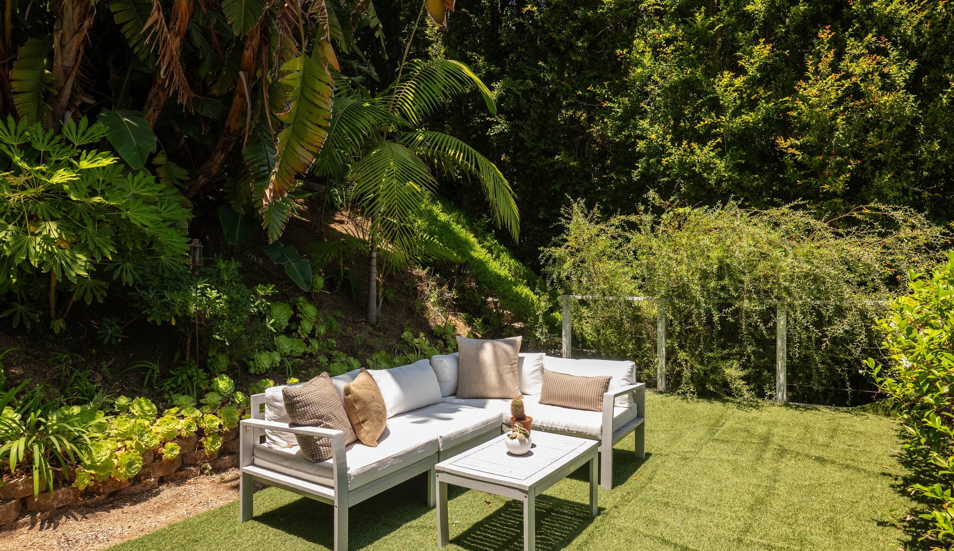 A turf lounge is placed among towering palms, tropical plants, and dense hillside landscaping, with outdoor seating arranged to take advantage of the quiet surroundings.
From there, a set of steps leads up to the next level of the backyard, where the atmosphere shifts from dining to relaxation. A manicured turf lounge is nestled among towering palms, tropical plants, and dense hillside landscaping, with outdoor seating arranged to take advantage of the quiet surroundings. Looking back from this elevated perch, you can appreciate how the terraced design layers each outdoor space, while a garden bridge disappears into the lush greenery above, adding another private corner to explore within the grounds.