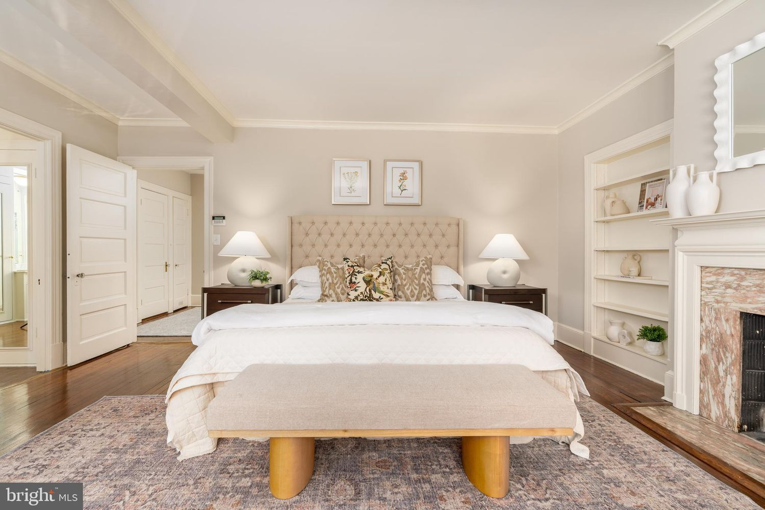The bedroom centers around a tufted upholstered bed, with hardwood floors, soft neutral walls, and crown molding reinforcing the home's traditional architecture.