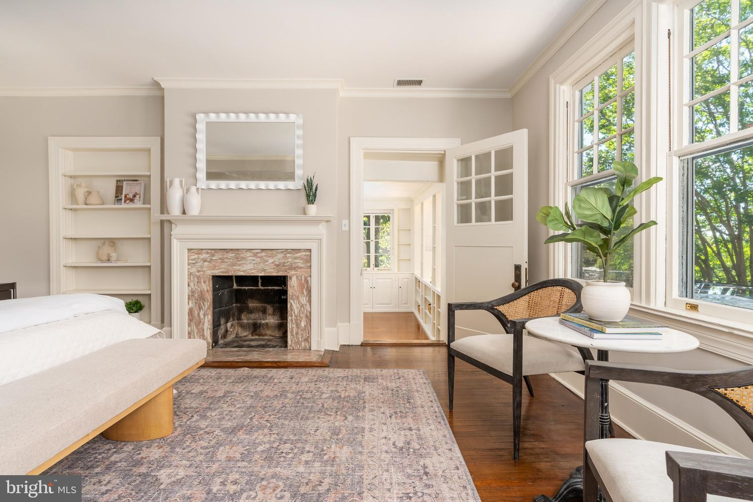 A marble fireplace with a detailed mantel sits on one wall of the room, framed by built-in shelving, while another features a small sitting area beneath the windows.