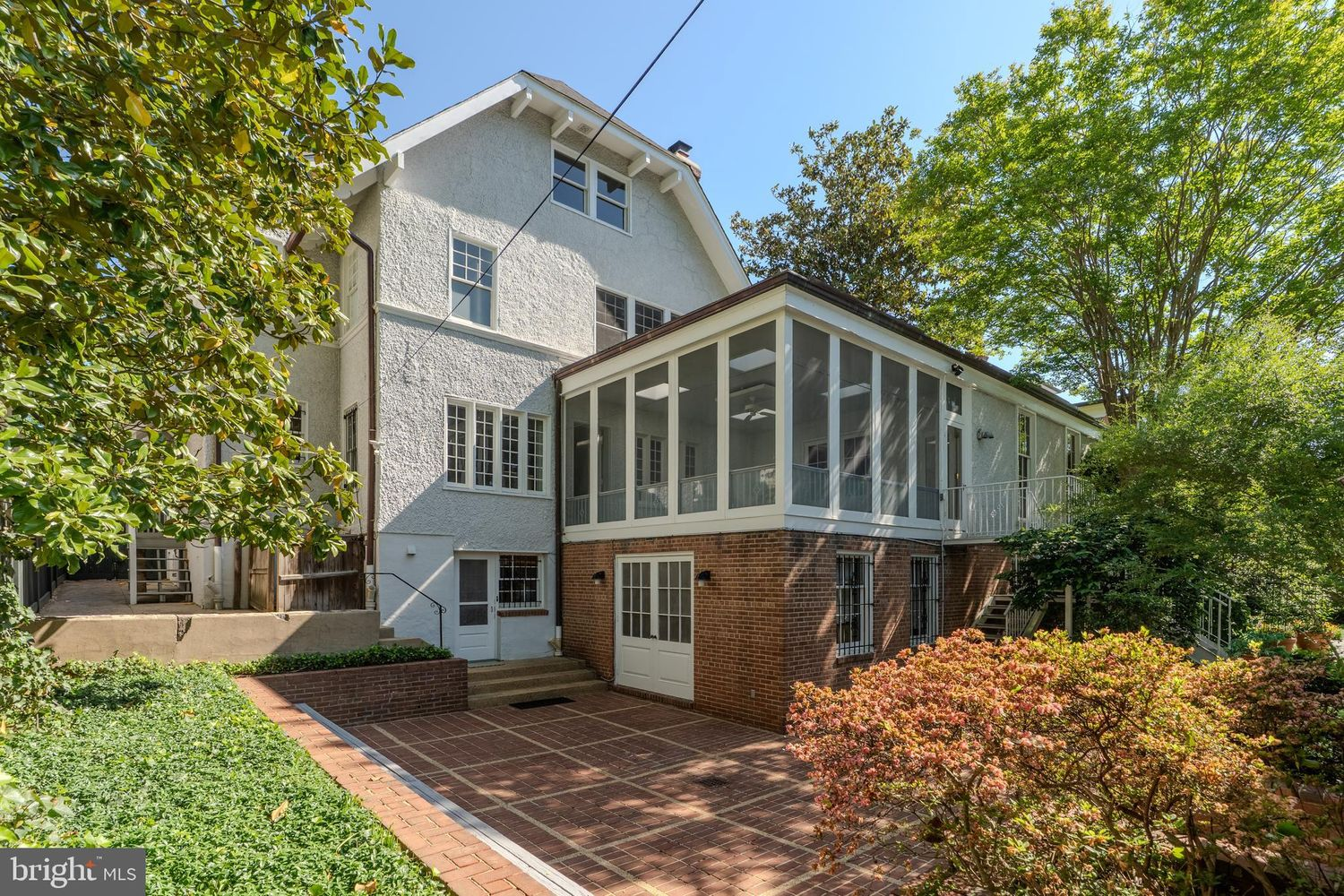 The back of the home opens onto a large brick patio tucked beneath the enclosed sunroom, creating a broad entertaining space partially shaded by the trees.