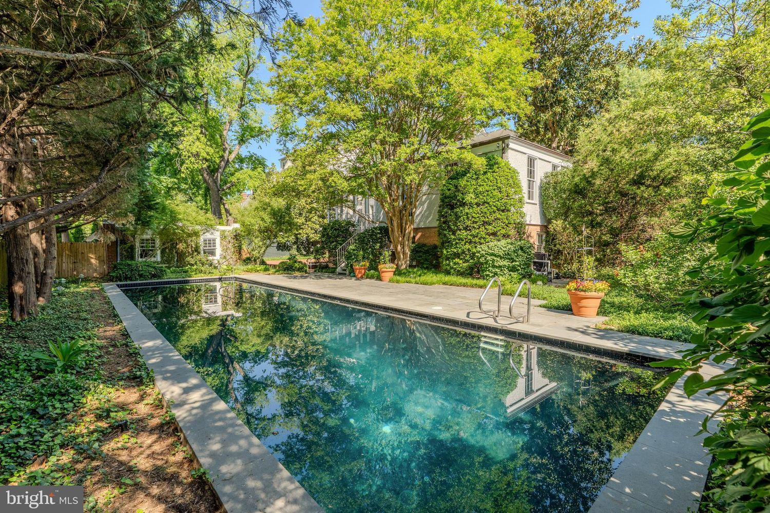A wide stone deck frames the length of the pool, leaving room for lounge seating, while expansive lawns, flowering planters, and towering trees surround the pool.