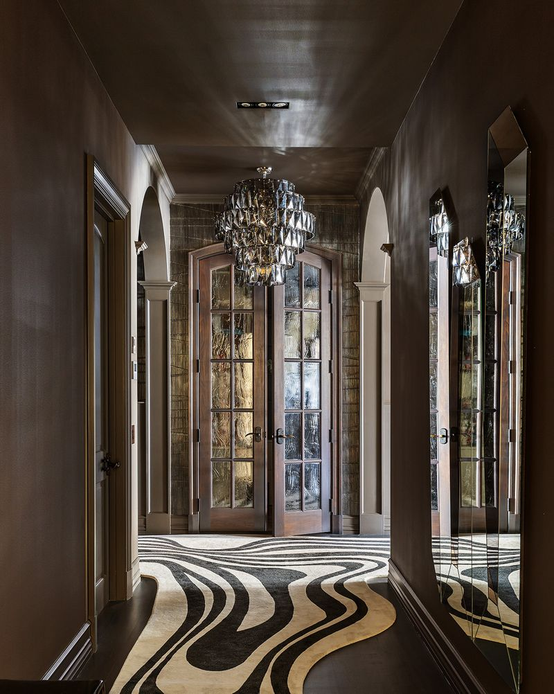 Rich, dark wall finishes, arched passageways, and oversized crystal chandeliers create a striking first impression, while sculptural mirrors and a flowing black-and-white runner add a contemporary edge.