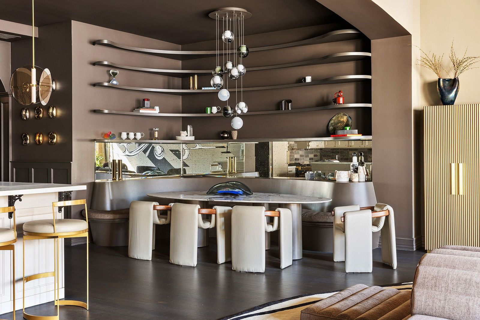 A marble-topped dining table sits beneath a cascading multi-globe chandelier, while mirrored detailing behind the banquette reflects the surrounding spaces.