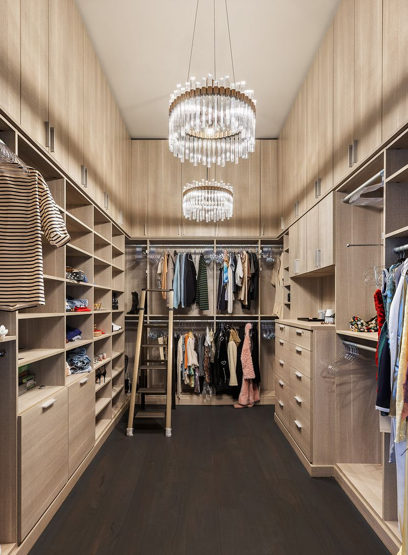 Floor-to-ceiling built-in cabinetry includes open shelving, hanging storage, and integrated drawers lining all sides of the room beneath two oversized crystal chandeliers.