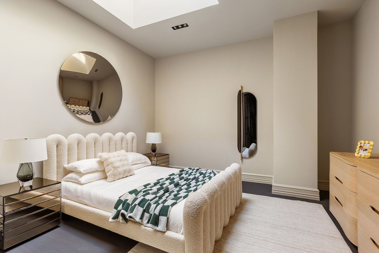 The guest bedroom features a sculptural upholstered bed beneath a large circular mirror that reflects light from the overhead skylight. 