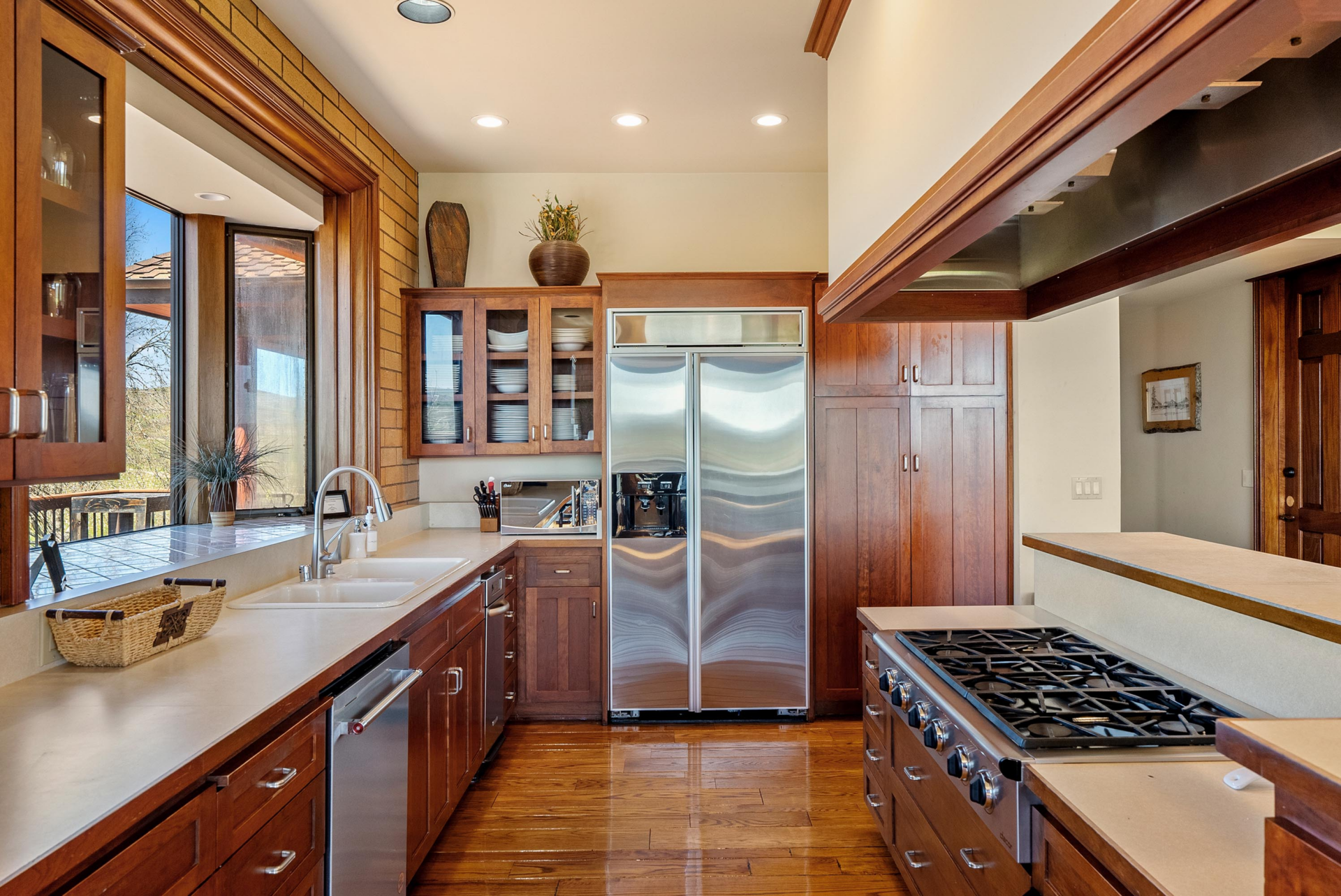 The kitchen extends into a narrower work zone lined with additional counters, two dishwashers, a stainless-steel refrigerator, and a professional-style range beneath the suspended hood.