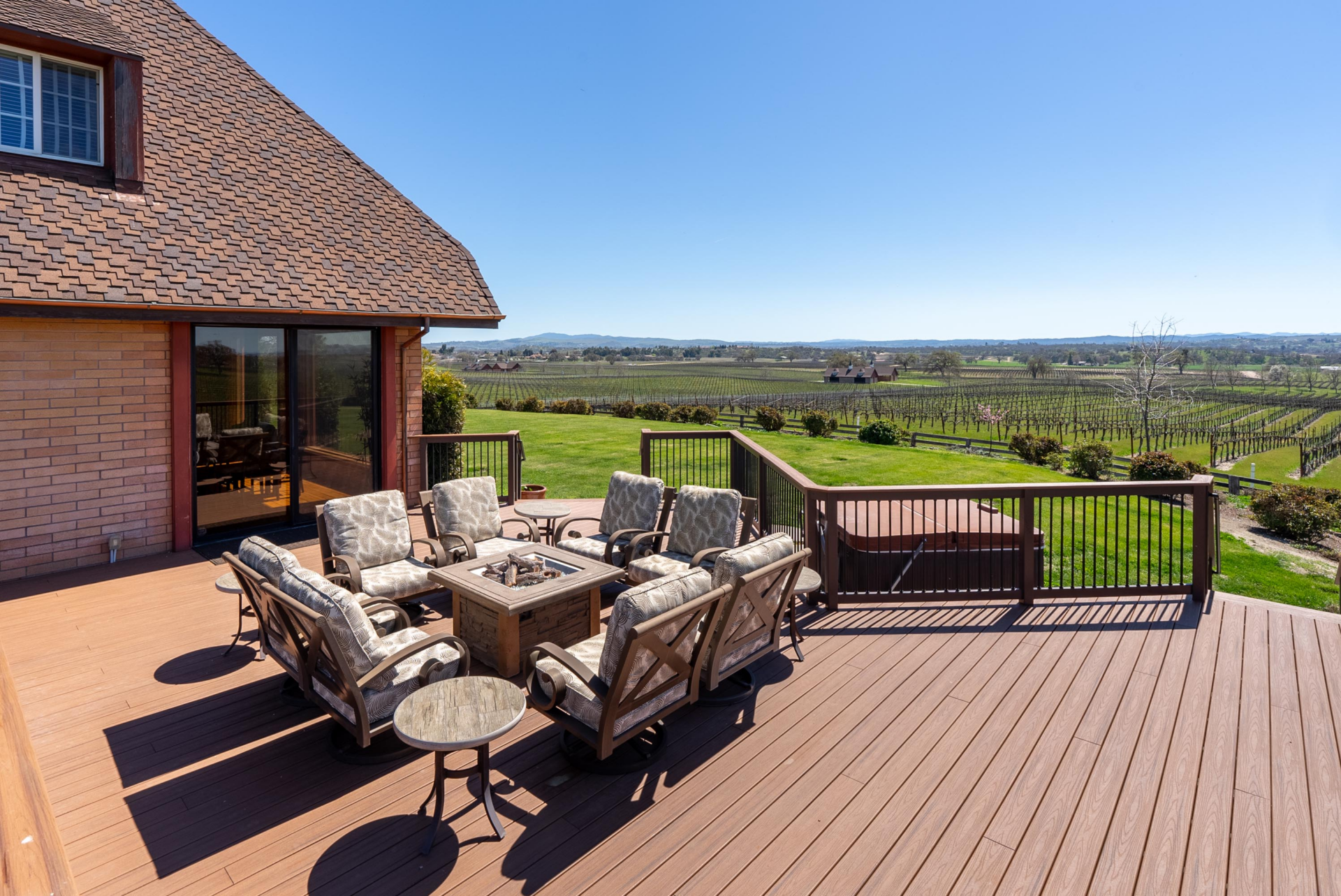 In the middle of the deck is a central seating area arranged around a square fire pit, with the railing kept low enough to preserve uninterrupted views across the lawn and surrounding vineyard rows.