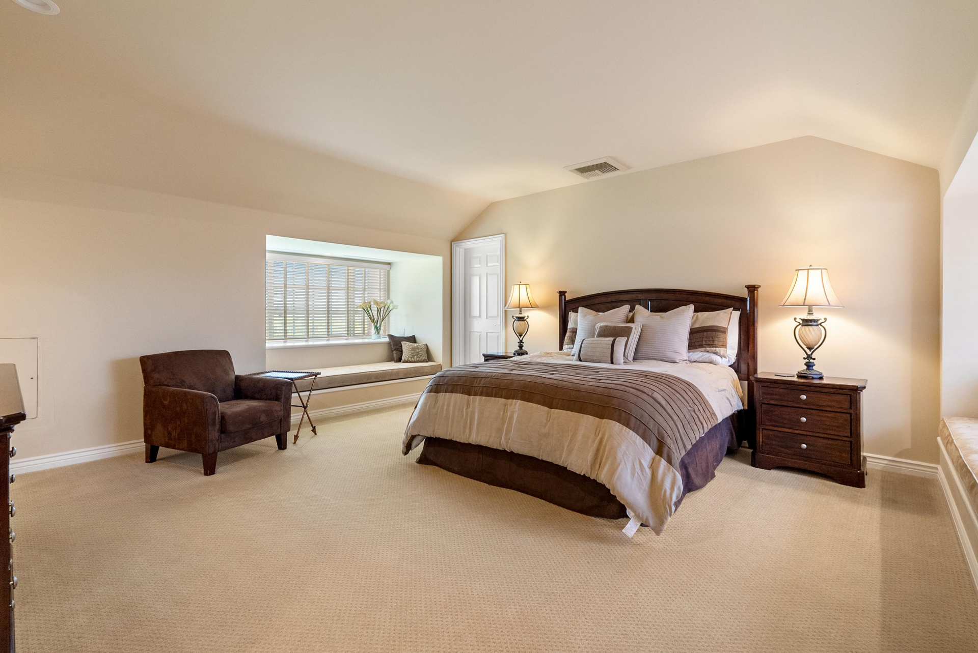 A larger bedroom is wrapped in soft neutral tones beneath a gently sloped ceiling, with the bed positioned along the room’s angled back wall. 