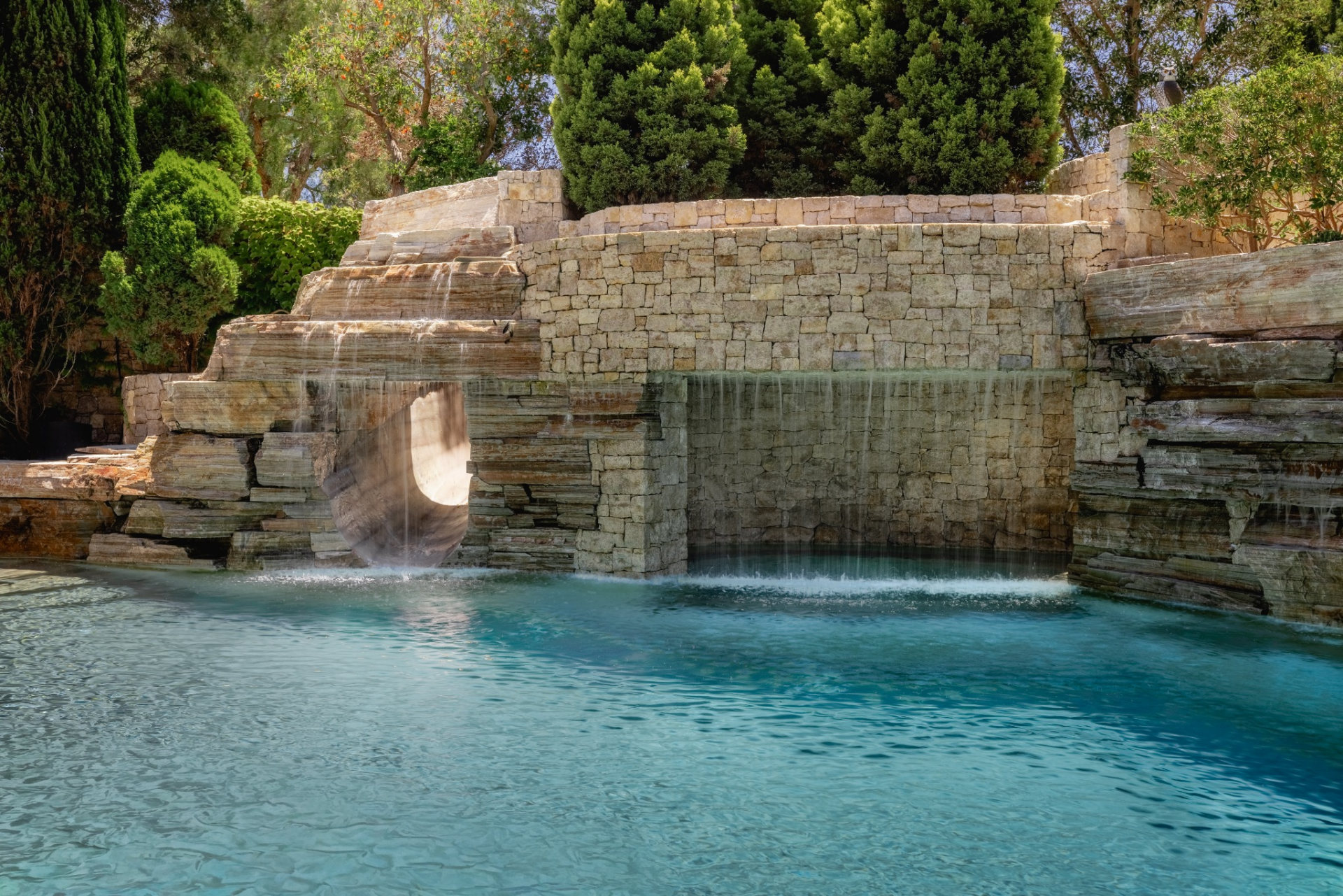 Layered rock walls rise along one side, where several waterfalls spill into the pool and a built-in slide curves through the stonework. 