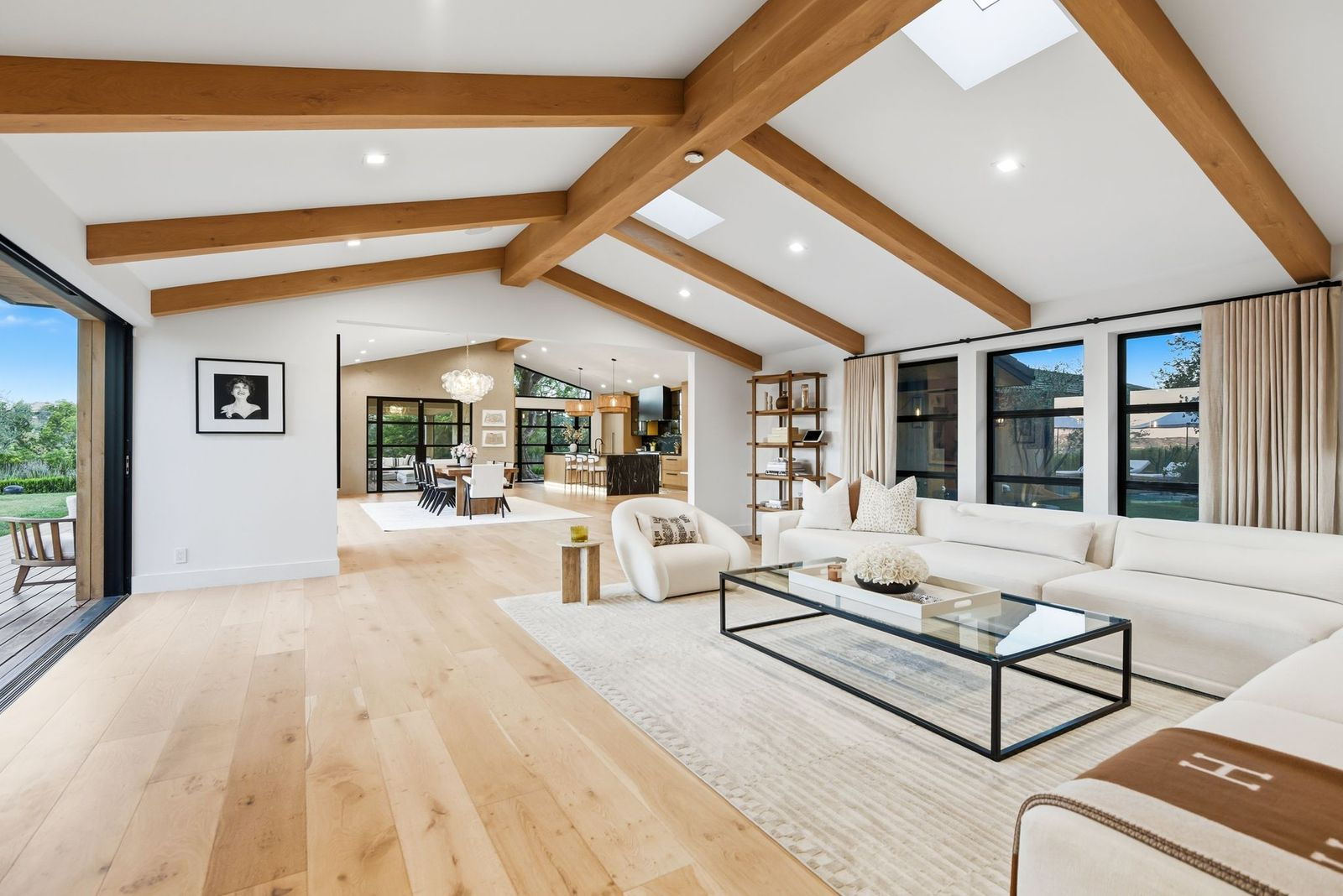 Exposed beams trace the pitched roofline above a long white sectional and low glass coffee table, while wide-plank wood floors continue toward the connected dining area and kitchen.