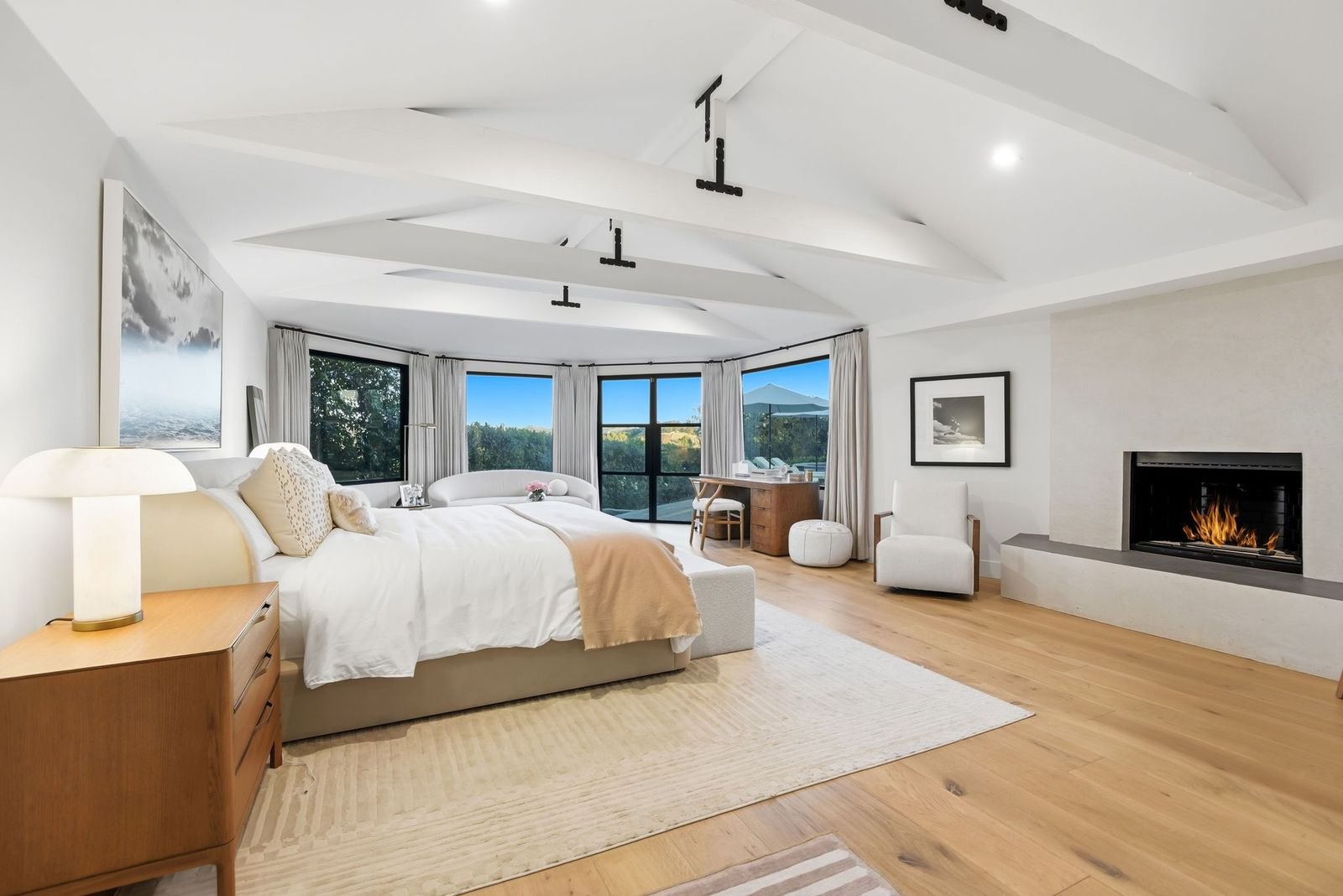 The primary bedroom opens beneath a vaulted ceiling with the bed occupying one side of the room, while a low fireplace and built-in hearth stretch across the opposite wall.
