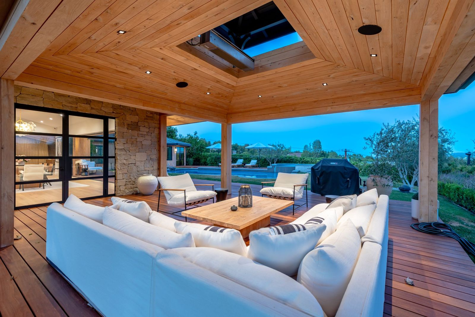 A large sectional and lounge chairs are arranged around a wood table, while stone walls and wide openings keep the space connected to both the pool and indoor living spaces.