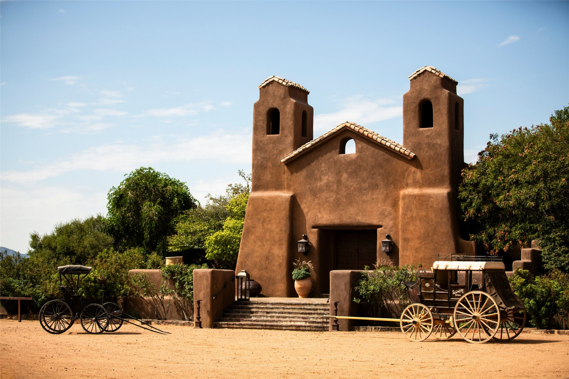 The chapel is marked by a simple adobe façade with twin bell towers, arched openings, and a short flight of stone steps leading to the entrance. 