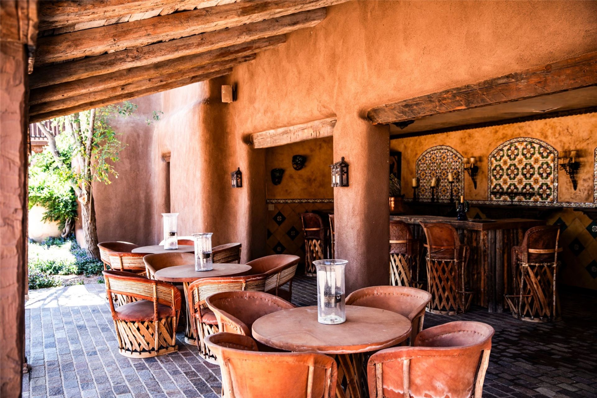 Beneath the covered section, round tables and leather chairs are arranged beside a built-in bar finished with rustic wood, patterned tile, and decorative metalwork.