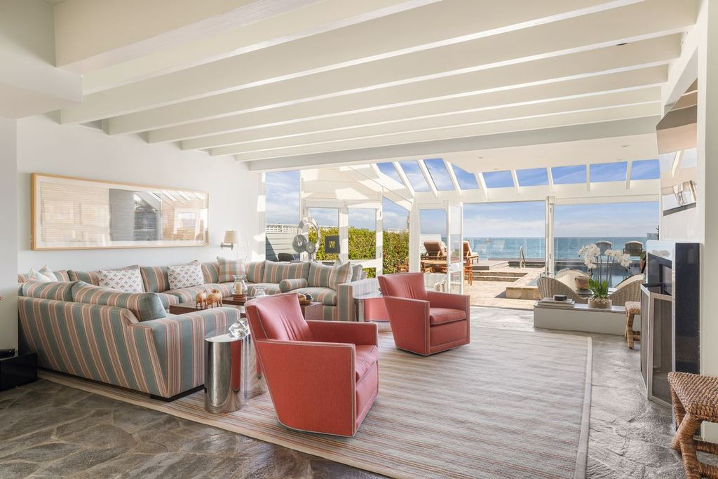 The room stretches beneath a white-painted beamed ceiling, with striped sofas and coral-toned lounge chairs arranged across a rug beside the fireplace. 